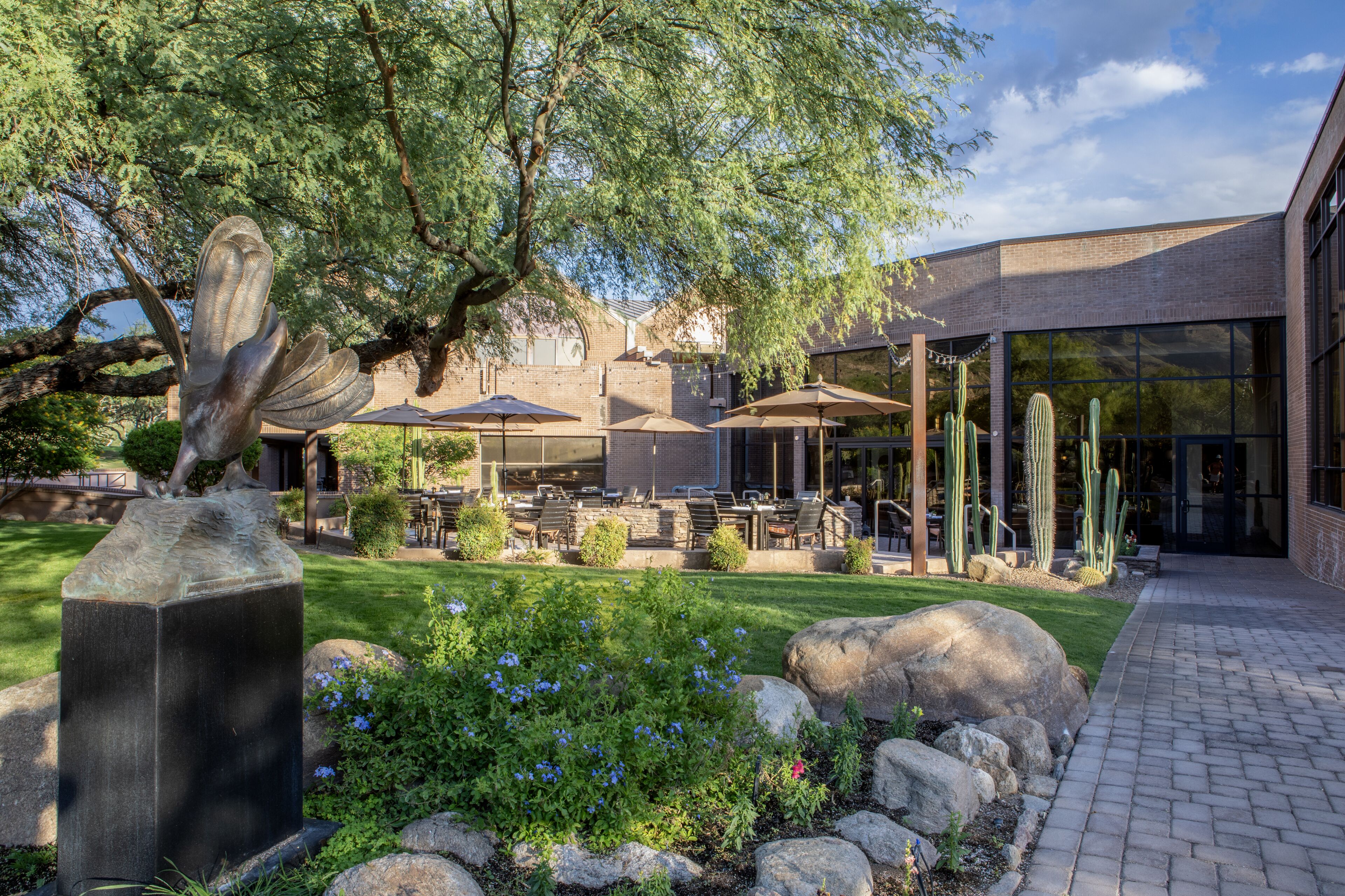 Une terrasse extérieure avec des tables et des parasols, entourée de cactus et d’une sculpture.