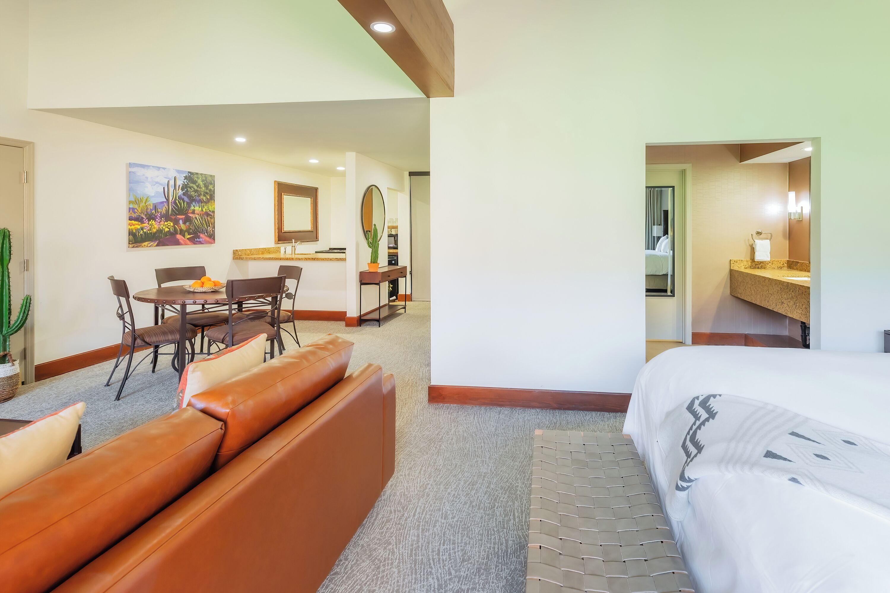 A hotel room with a bed, a sofa, a dining table with chairs, a small kitchen area, and a bathroom visible through an open door.
