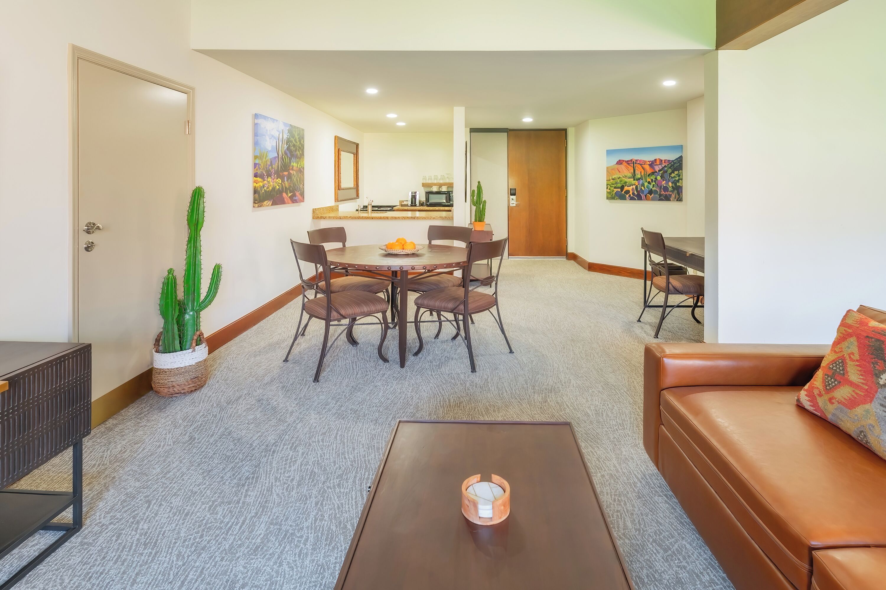 A dining area with a round table, four chairs, a coffee table, and a sofa. There are two potted cacti on the floor.