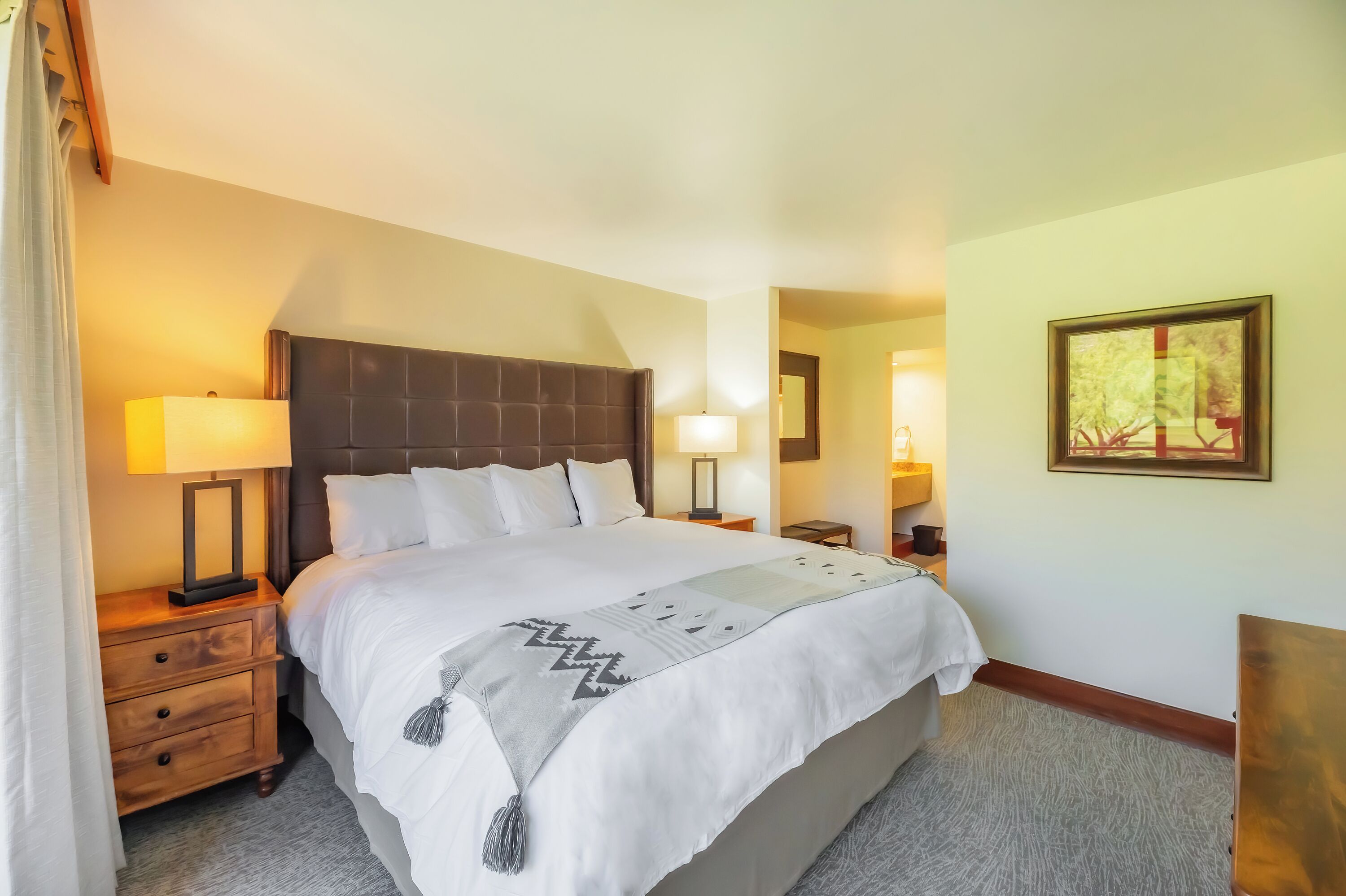 A neatly made bed with a headboard, two bedside tables with lamps, a framed picture on the wall, and a carpeted floor.