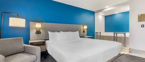A hotel room with a large bed, a grey armchair, a nightstand, and a mirror.