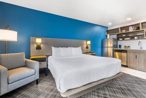 Habitación de hotel con una cama grande, un sillón gris, una mesita de noche con lámpara, una pequeña mesa con lámpara, una cocina integrada con refrigerador y una pared de acento azul.