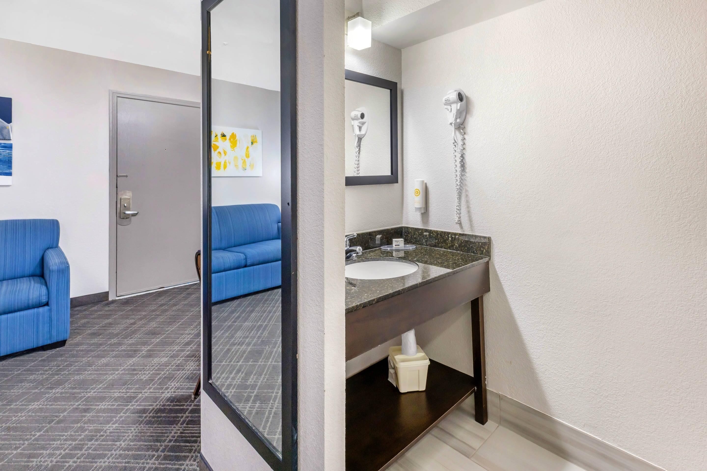 Baño de hotel con lavamanos, espejo y secador de pelo. Se ve una silla azul y una puerta al fondo.