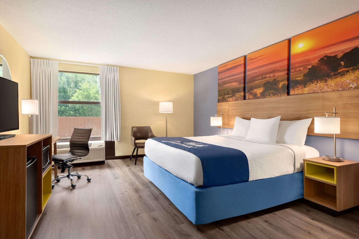 A hotel room with a bed, desk, chair, and a painting of a sunset.