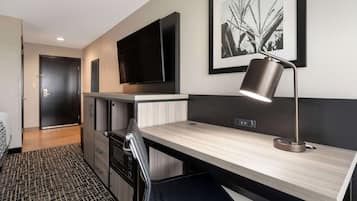 A modern hotel room with a wooden desk, a black chair, a flat-screen TV mounted on the wall, a framed picture, and a desk lamp.