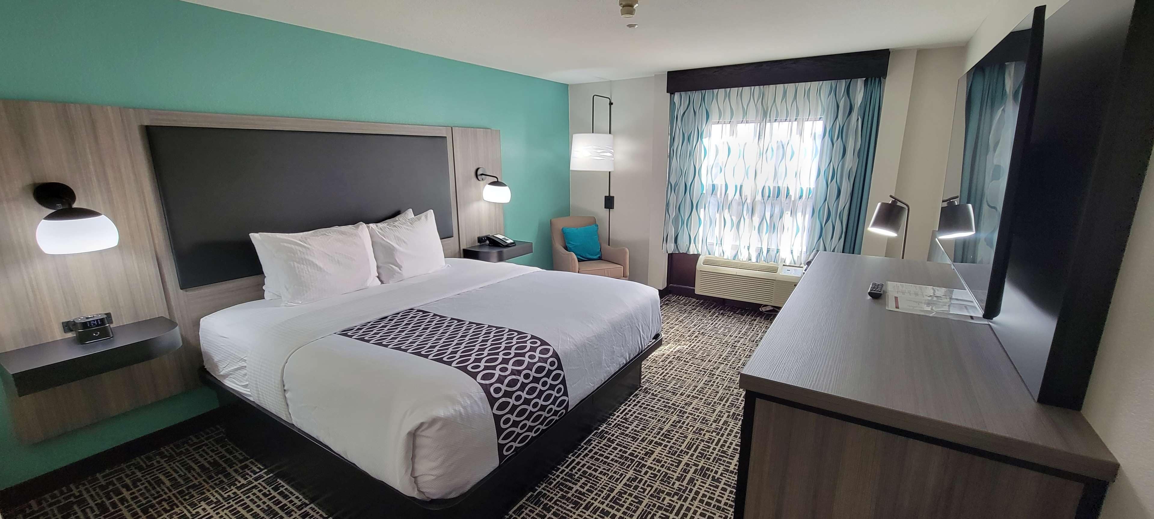 Une chambre d’hôtel avec un grand lit, des tables de chevet, un bureau, une chaise, une grande fenêtre avec des rideaux et un téléviseur à écran plat.