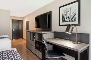 Una habitación de hotel moderna con una cama, un escritorio con silla, un microondas, un televisor de pantalla plana y un cuadro en la pared.