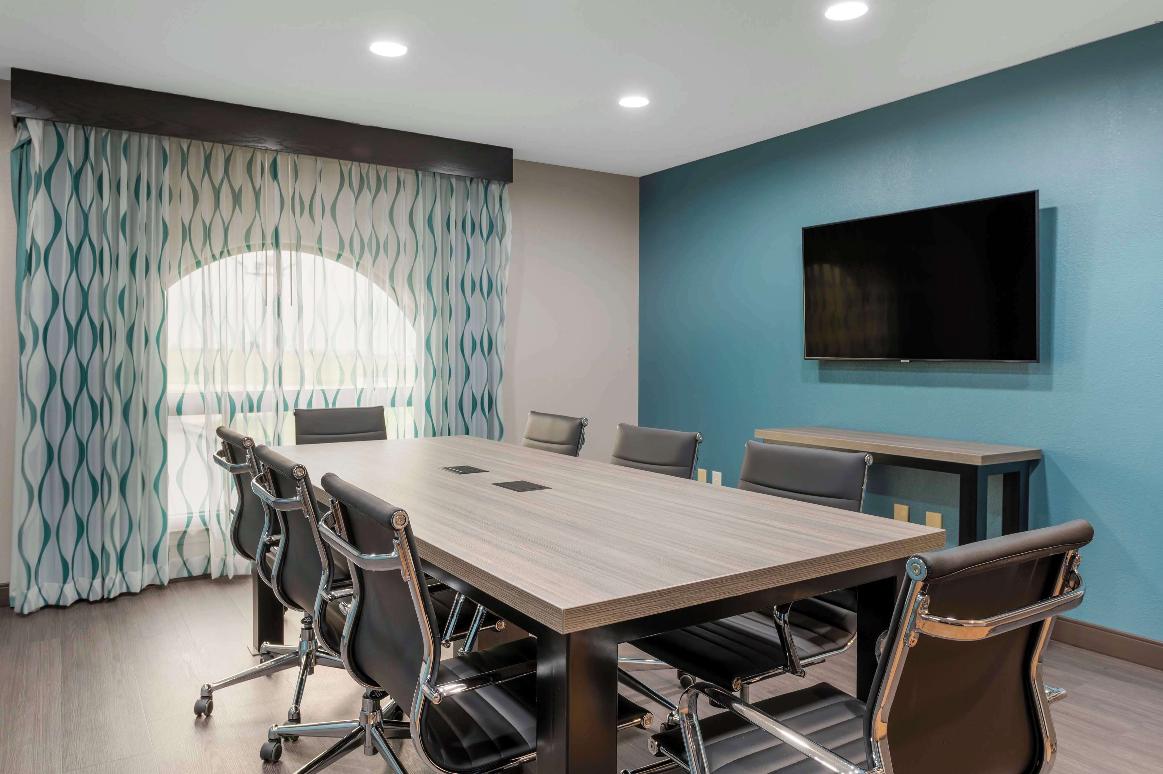 Uma sala de conferências com uma mesa comprida, seis cadeiras, uma TV fixada na parede e uma janela com cortinas estampadas.