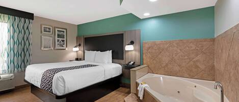 A hotel room with a bed, a bedside table with a phone, a wall-mounted lamp, a large bathtub, and a wall-mounted mirror.