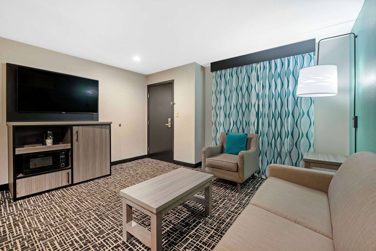 A modern hotel room with a flat-screen TV, kitchenette, sofa, and patterned carpet.