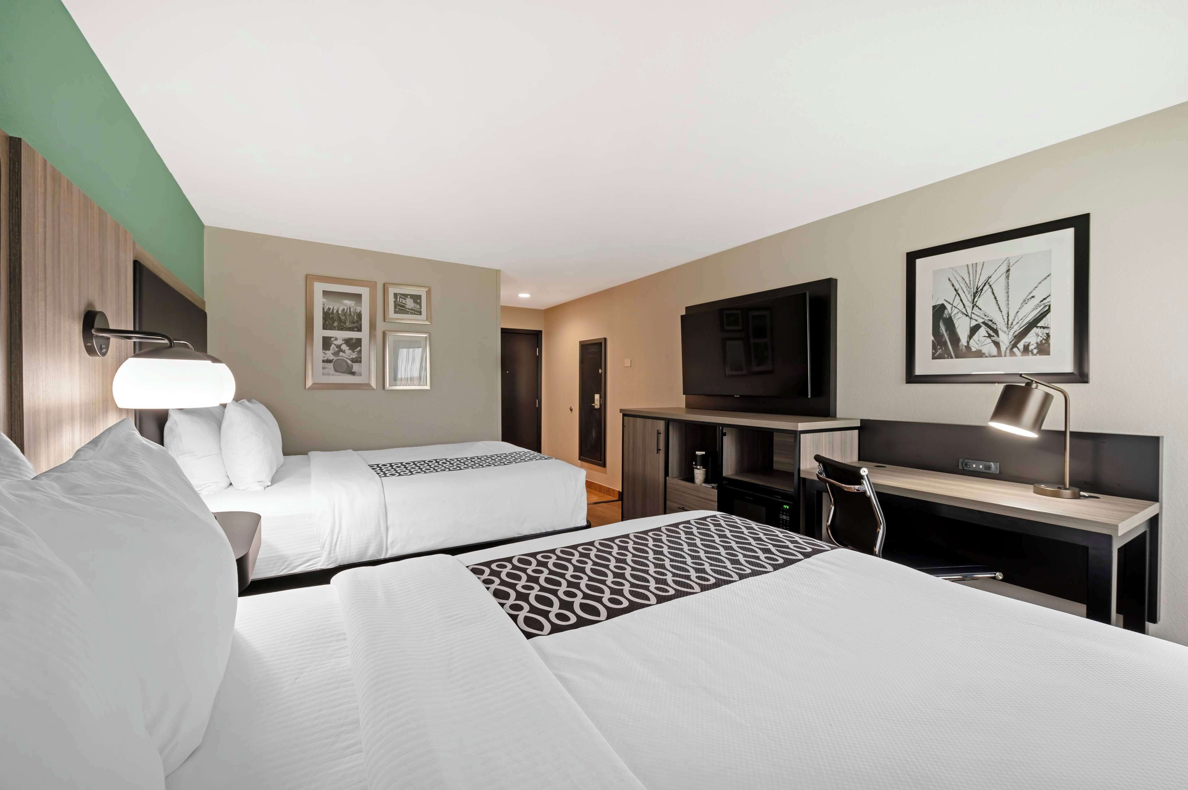 Ein Hotelzimmer mit zwei Betten, einem Schreibtisch, einem Fernseher und gerahmten Bildern an der Wand.