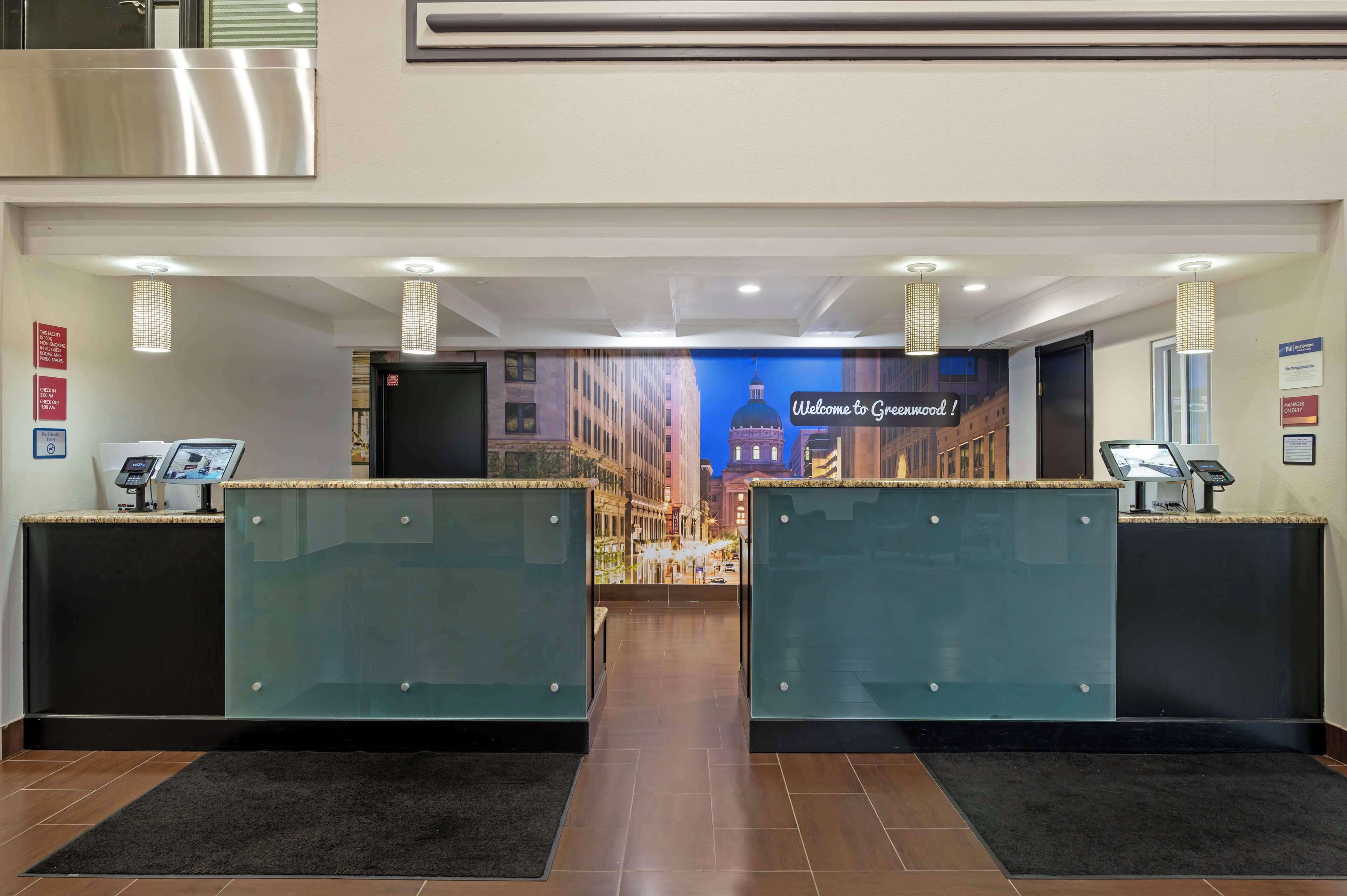 A hotel reception area with two glass-enclosed counters, a digital check-in kiosk, and a welcome sign.