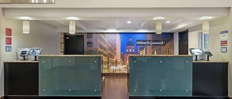 A hotel reception area with two glass-enclosed counters, a digital check-in kiosk, and a welcome sign.