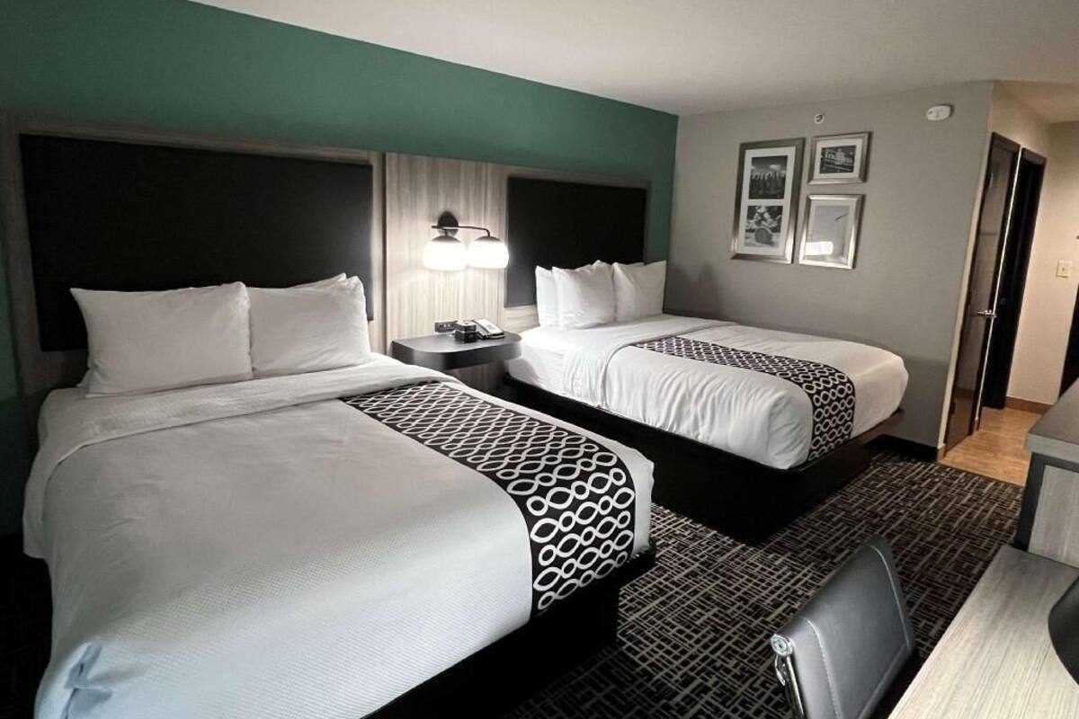A hotel room with two beds, a desk, a chair, a lamp, and framed pictures on the wall.