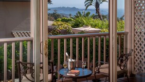 A balcony with a table, chairs, and a bottle of wine, overlooking a lush garden and a scenic view of the ocean.
