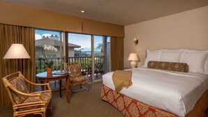 A hotel room with a large bed, a chair, a table, and a view of a balcony with a palm tree.