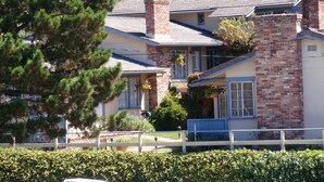 A two-story house with a brick chimney, a balcony with hanging flower baskets, and a well-maintained front yard with hedges.