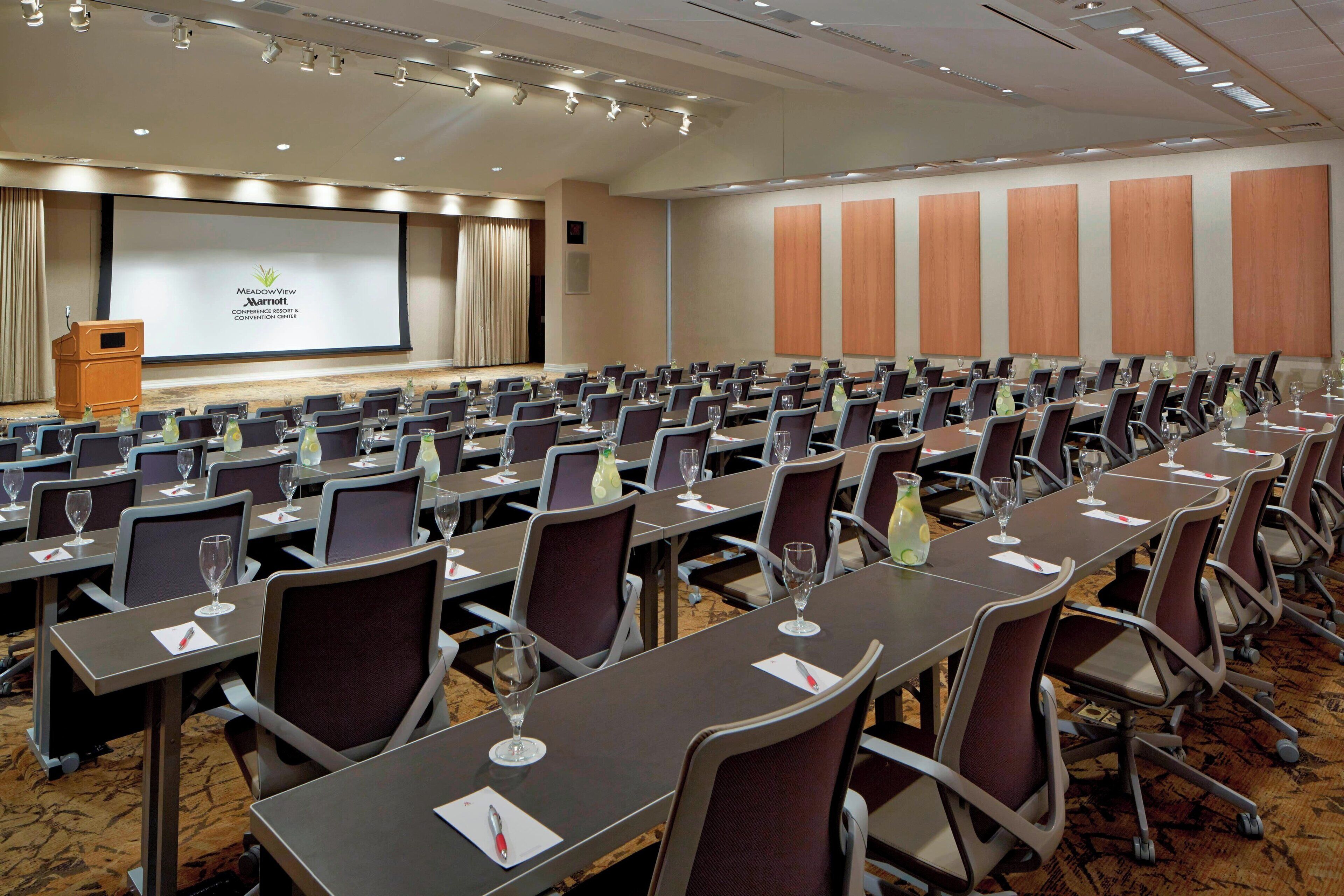 Un salón de conferencias con filas de mesas, sillas y una pantalla para presentaciones al frente.
