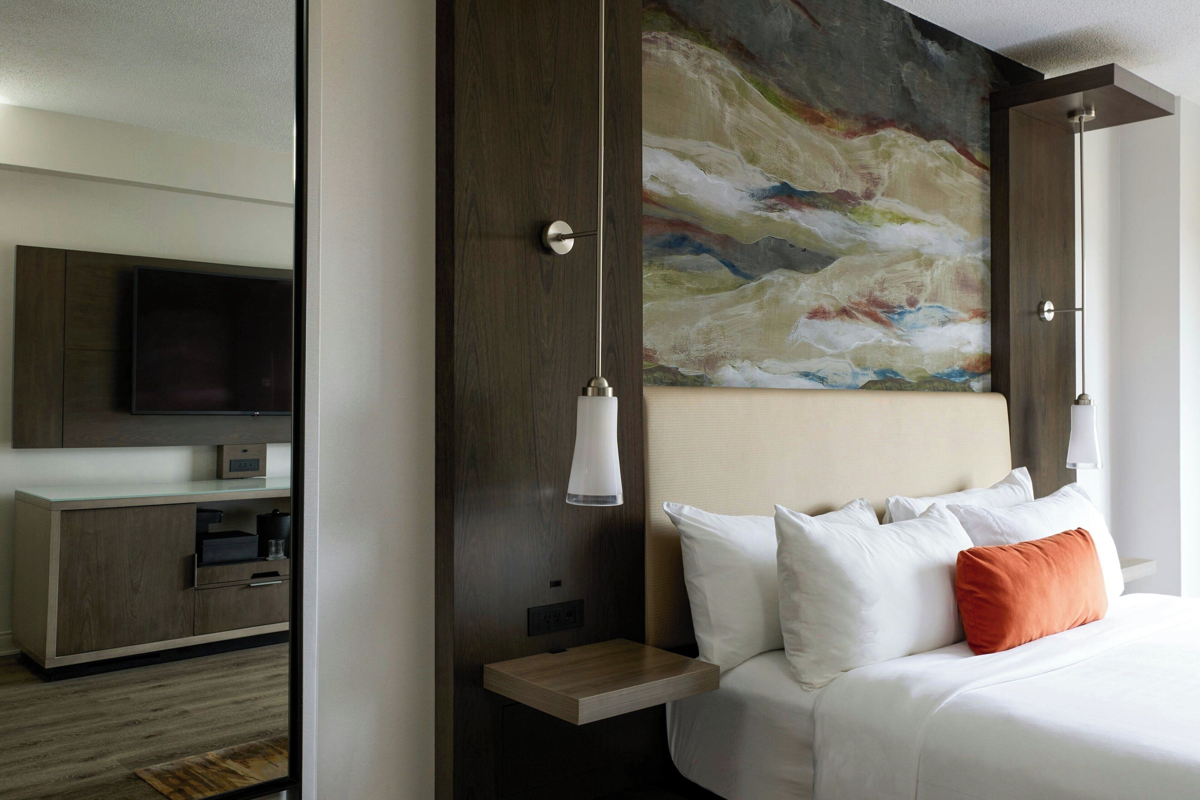 A hotel room with a bed, a nightstand, a TV, a painting, and a wooden cabinet.