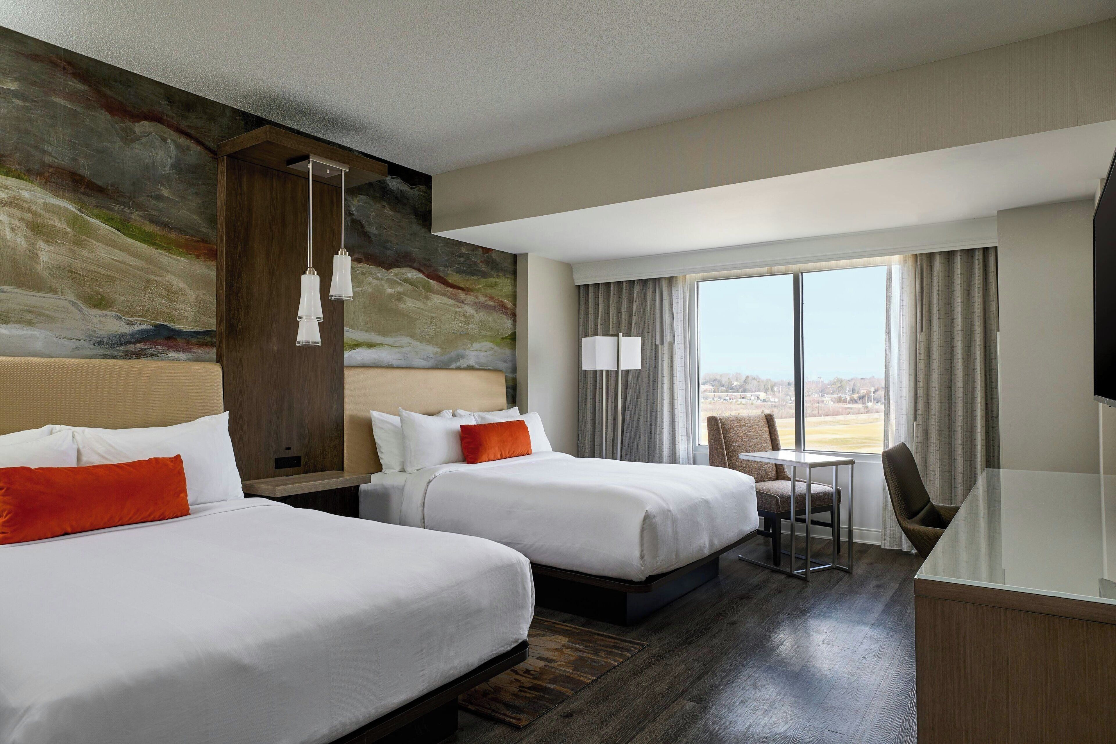A hotel room with two beds, a desk, a chair, a TV, and a large window with a view.