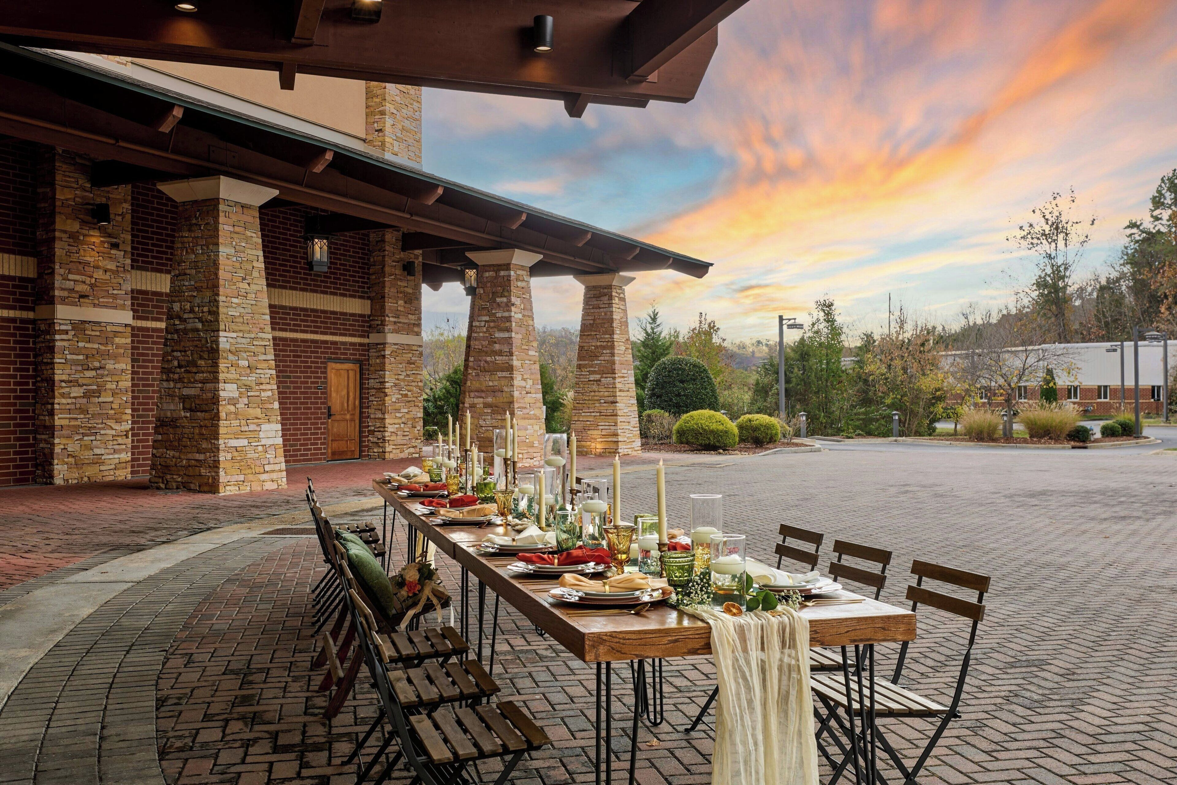 An outdoor dining area with a brick building, a long table set for a meal, and a sunset in the background.