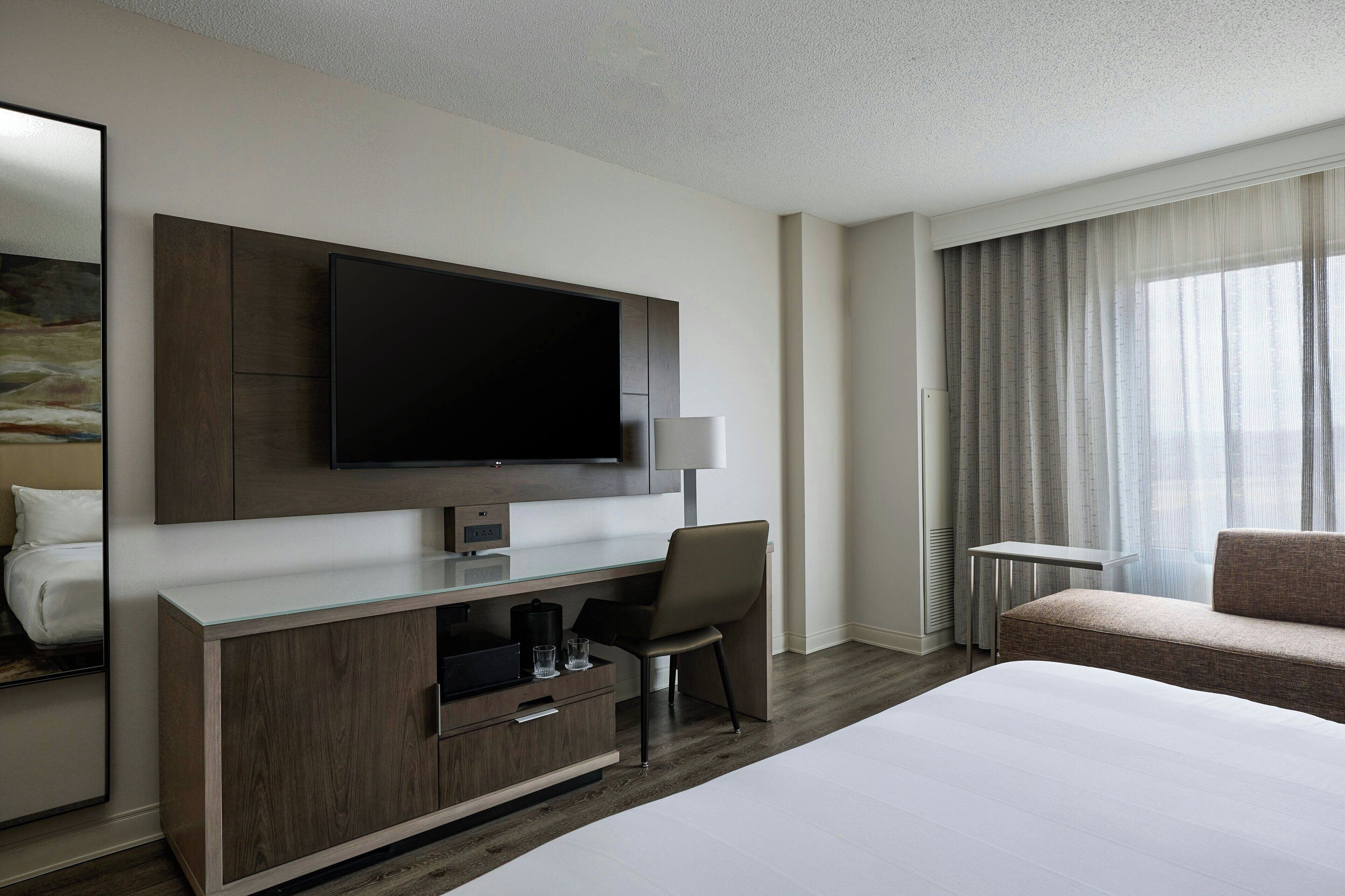 A modern hotel room with a large bed, a flat-screen TV mounted on a wooden cabinet, a desk with a chair, and a sofa.
