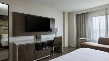 A modern hotel room with a large bed, a flat-screen TV mounted on a wooden cabinet, a desk with a chair, and a sofa.