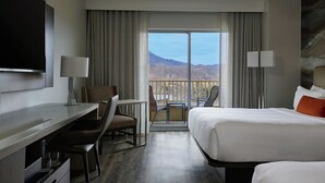 A hotel room with a bed, a desk, a chair, a TV, and a balcony with a view of the mountains.