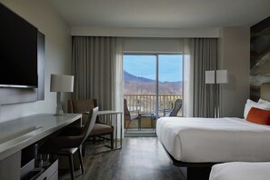 A hotel room with a bed, a desk, a chair, a TV, and a balcony with a view of the mountains.