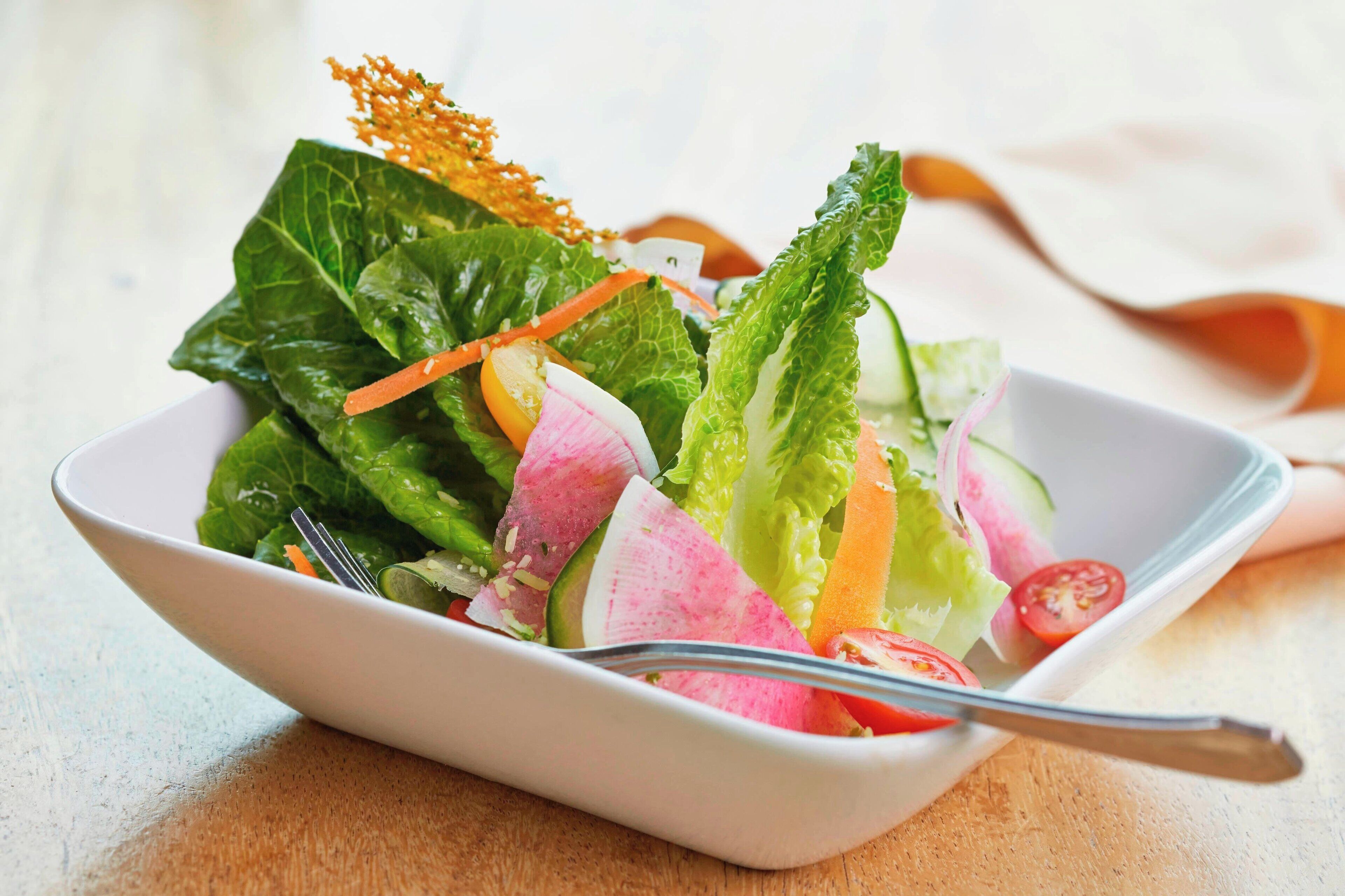 A white bowl containing a salad with lettuce, radishes, cucumber, and carrot slices.