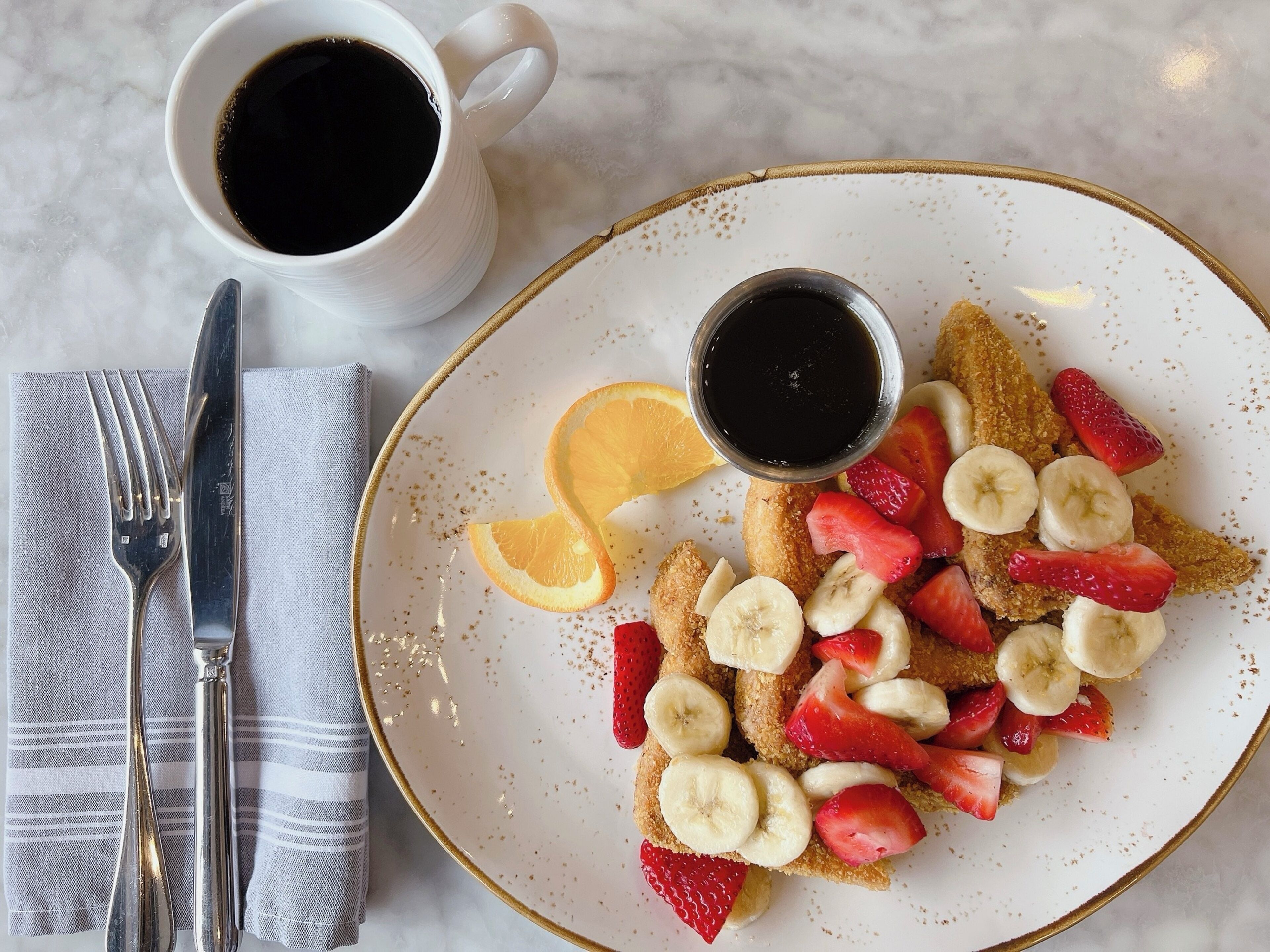 A breakfast plate with fruit and syrup, a cup of coffee, and cutlery on a marble surface.