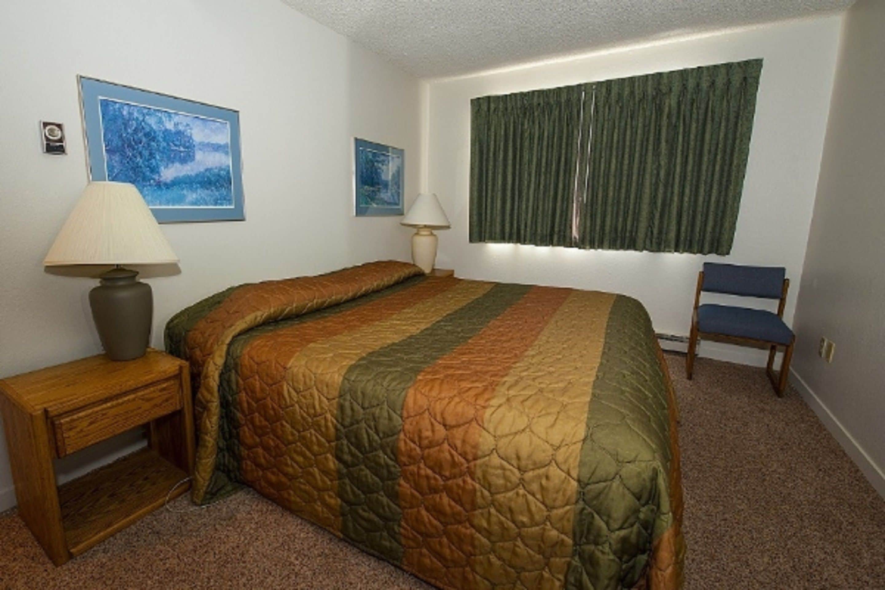 A bedroom with a bed, two bedside tables, a chair, a window with curtains, and two framed pictures on the wall.