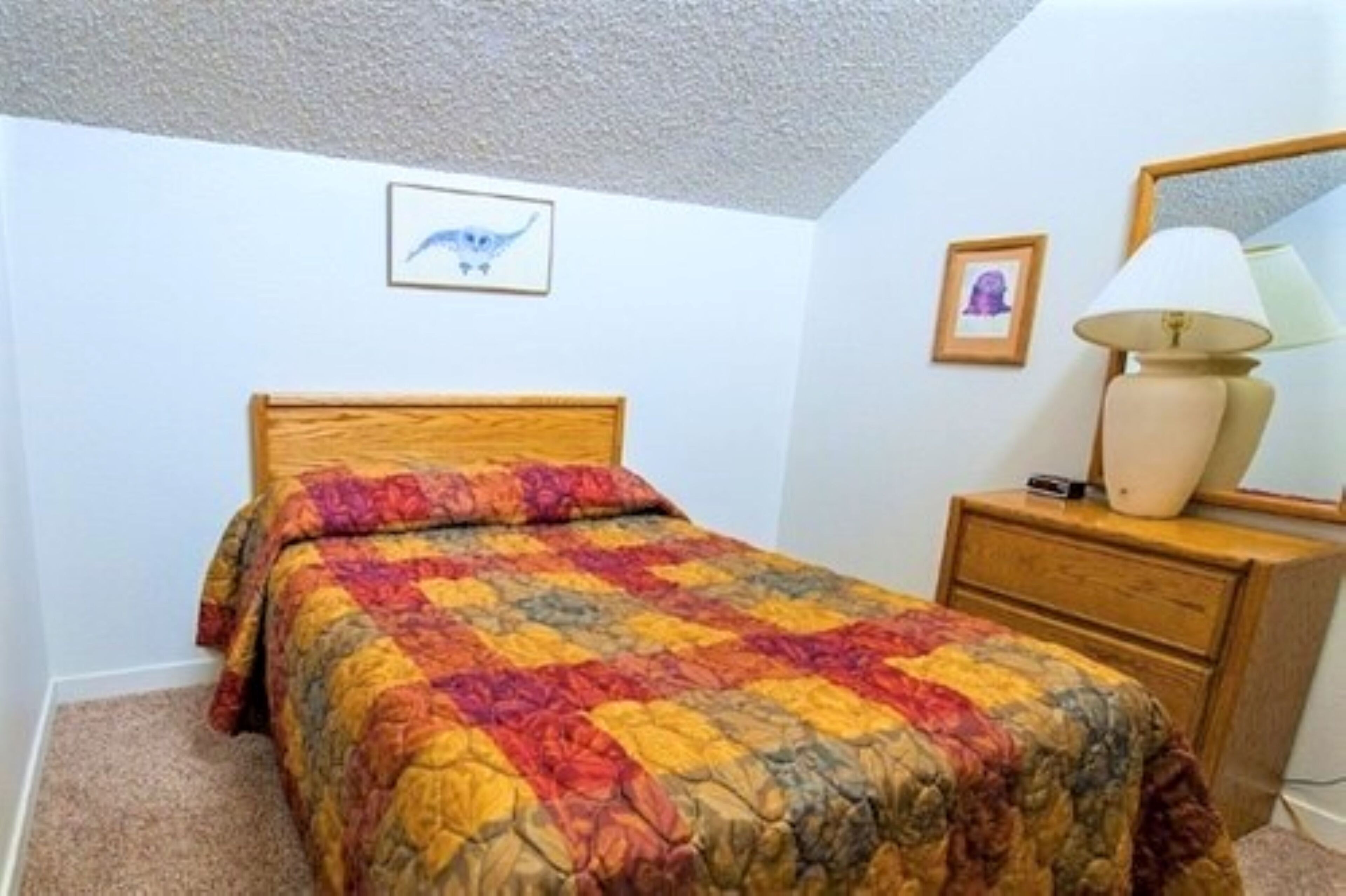 A bedroom with a wooden bed, colorful quilt, a bedside lamp, a dresser, and a framed picture on the wall.