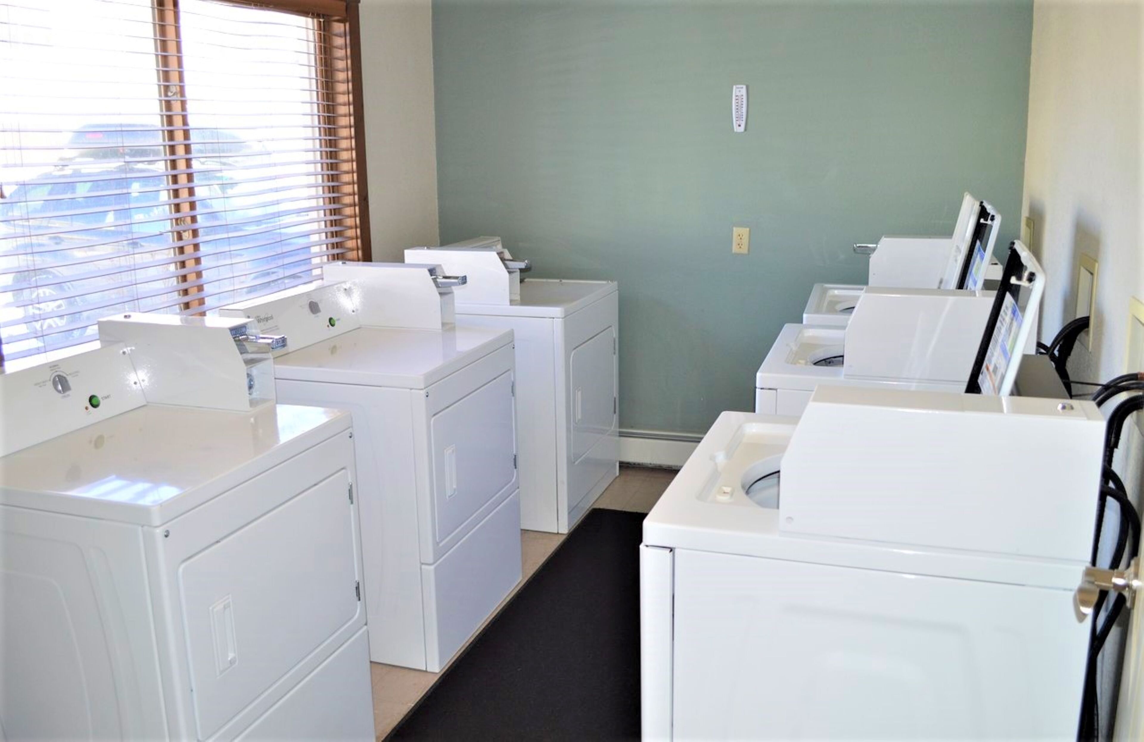 A laundry room with multiple white washing machines and dryers, a black mat on the floor, and a window with blinds.