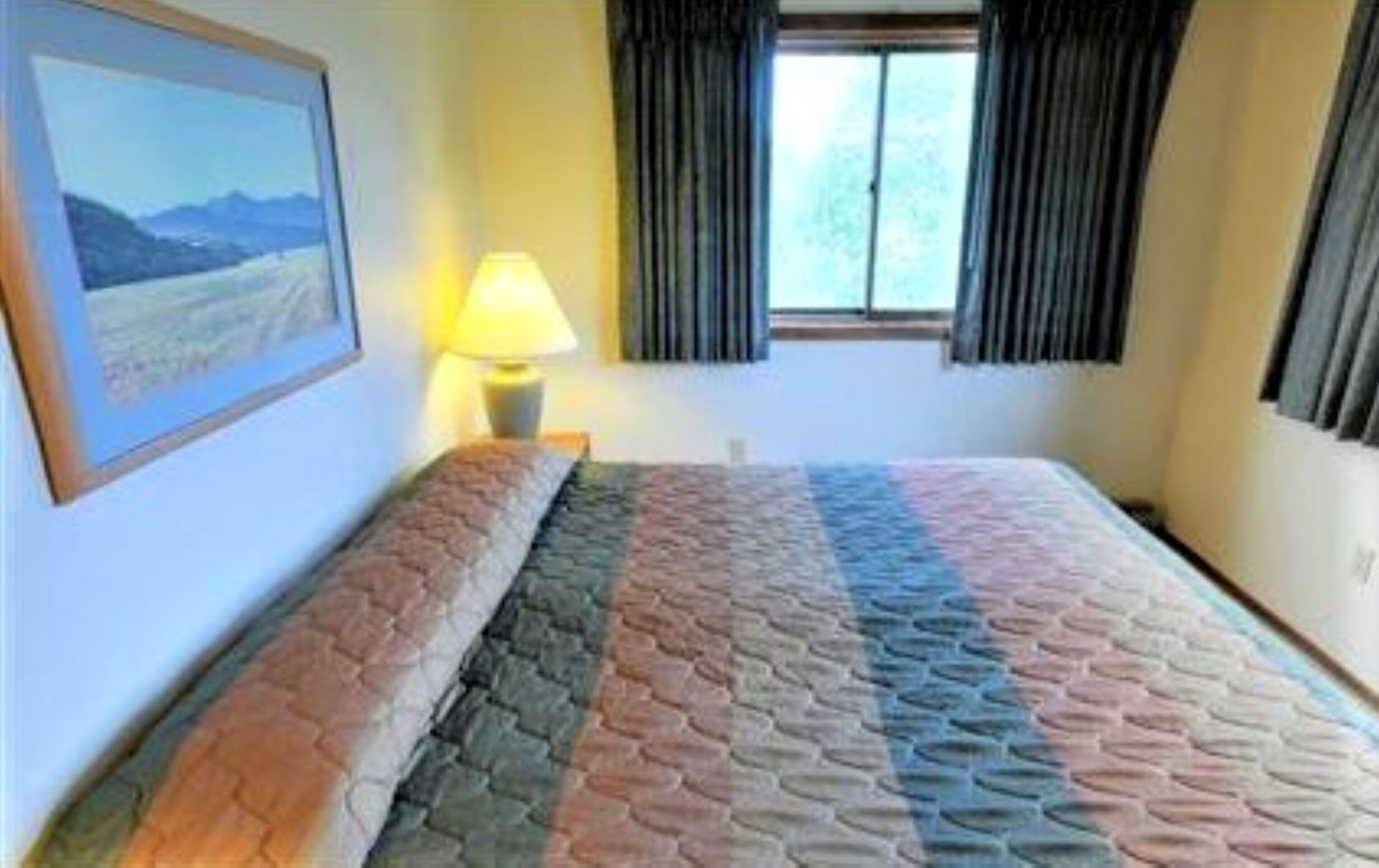 Une chambre d’hôtel avec un lit, une fenêtre avec des rideaux, une lampe et un tableau encadré au mur.