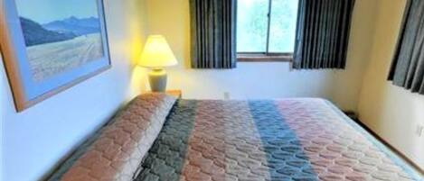 Une chambre d’hôtel avec un lit, une fenêtre avec des rideaux, une lampe et un tableau encadré au mur.