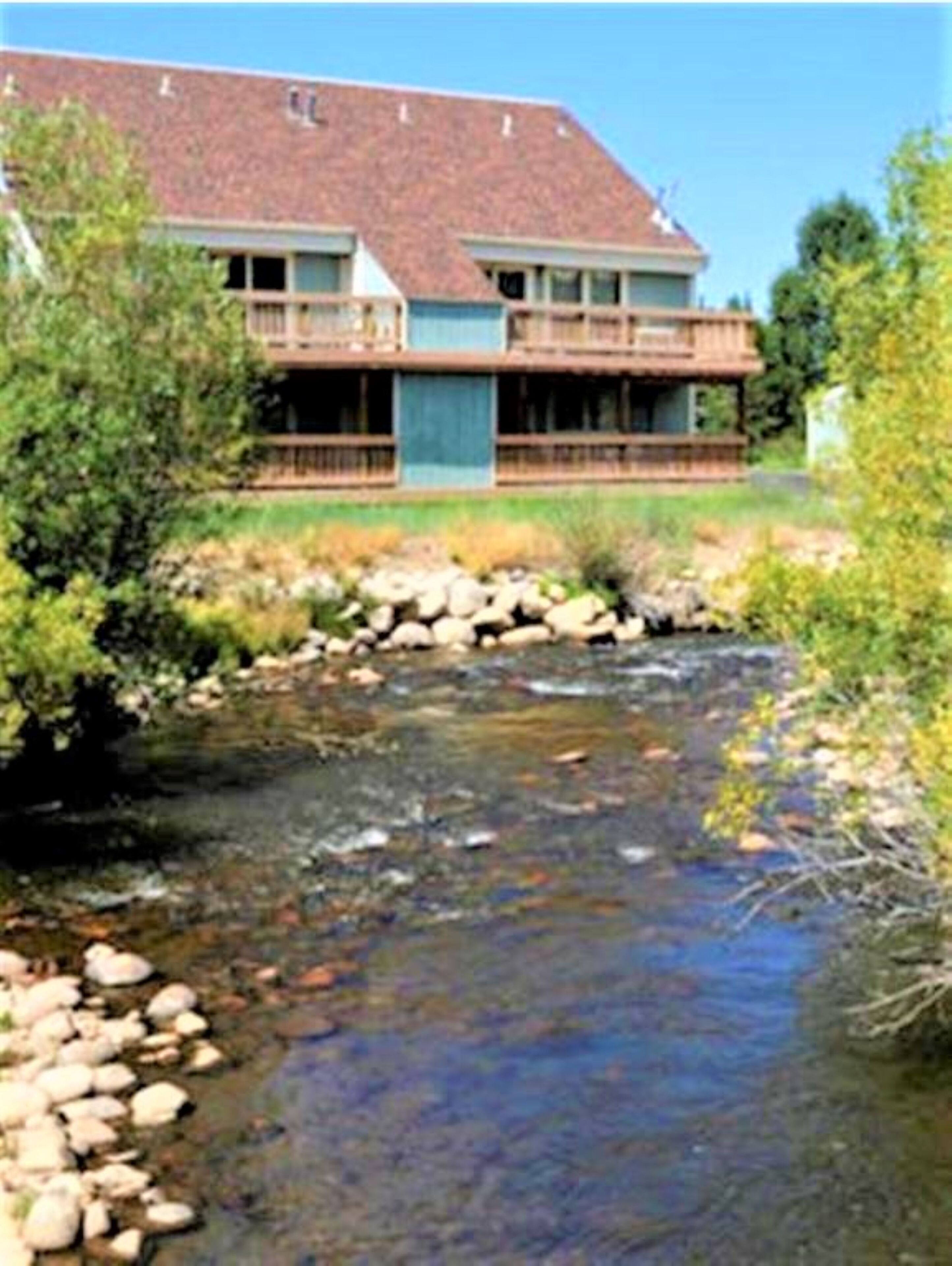 A two-story house with a stone foundation, a wooden deck, and a small stream in front.