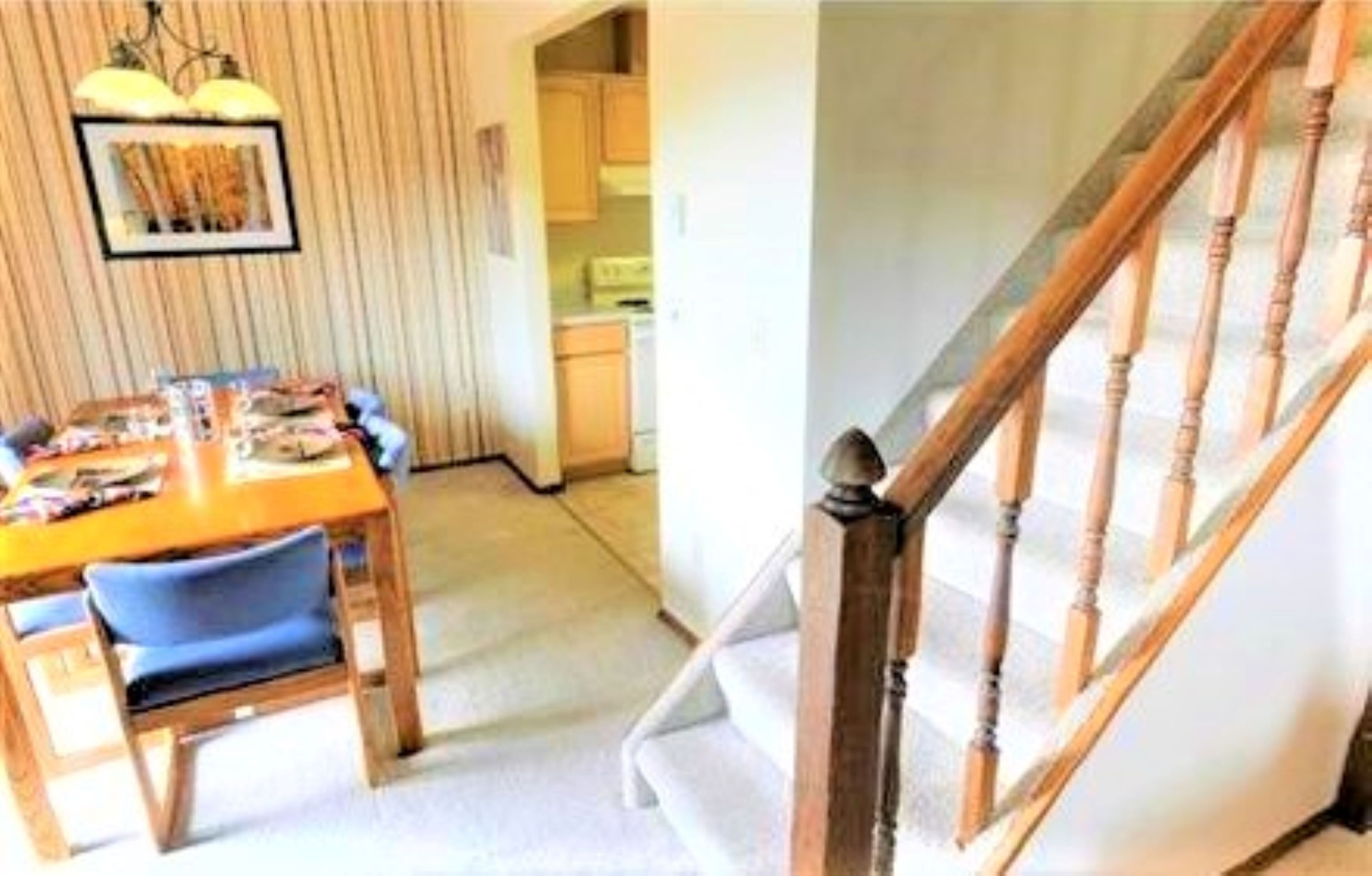 A dining area with a wooden table and chairs, a kitchen in the background, and a wooden staircase.