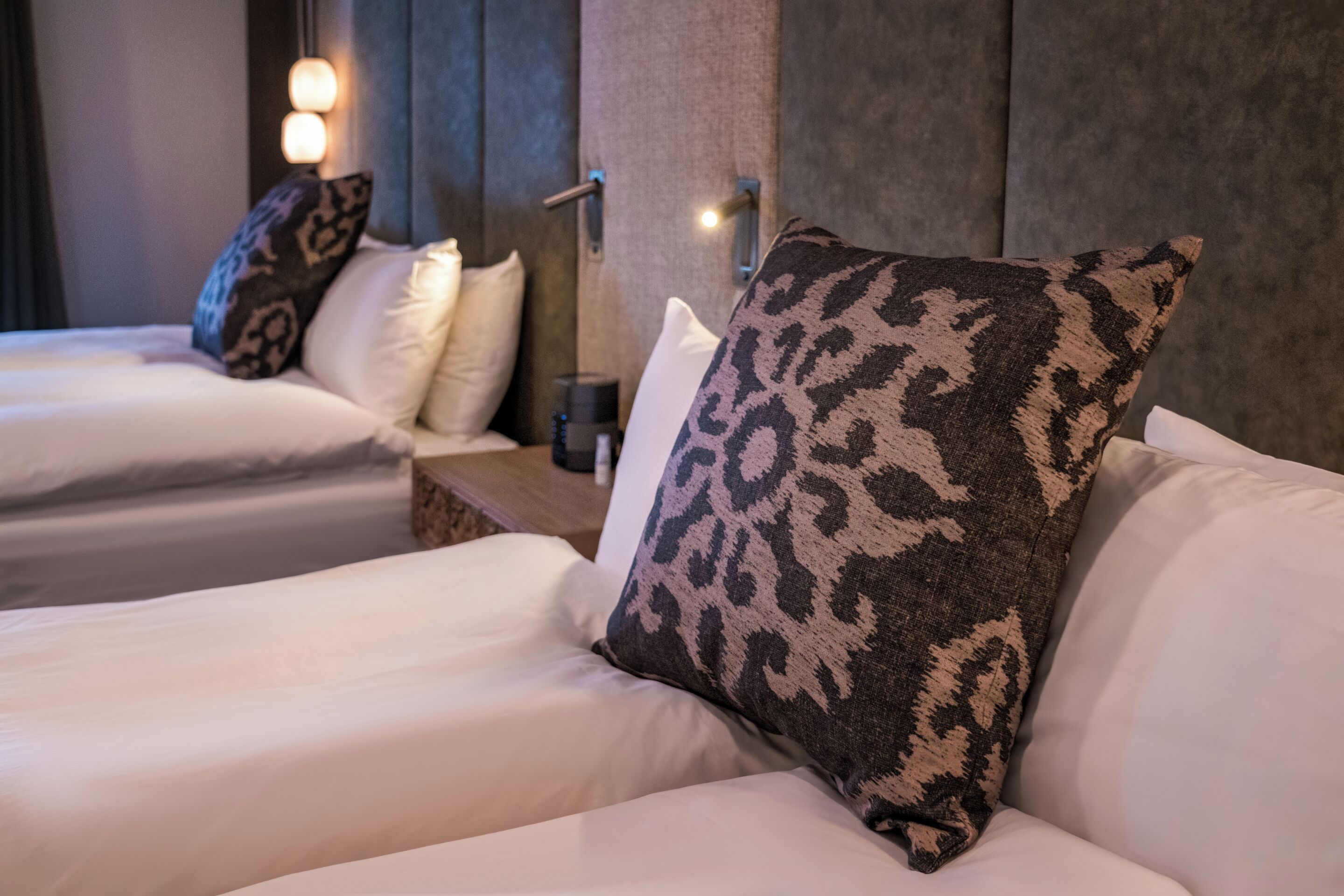 A hotel room with two beds, each with white bedding and patterned pillows. There are bedside tables with lamps and a wall-mounted light.