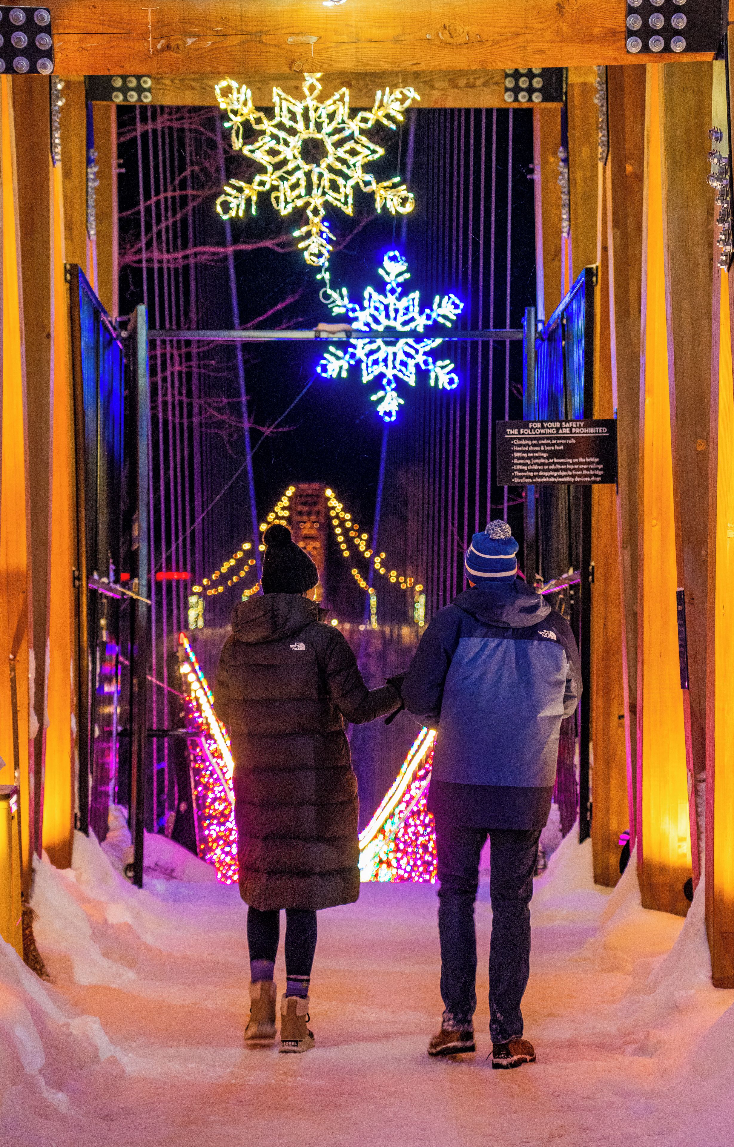Two people walking on a snow-covered bridge with illuminated snowflake decorations.