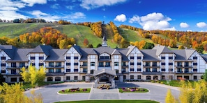 Um hotel grande com vários andares, um pátio central e vista para uma paisagem montanhosa coberta por folhagem outonal.