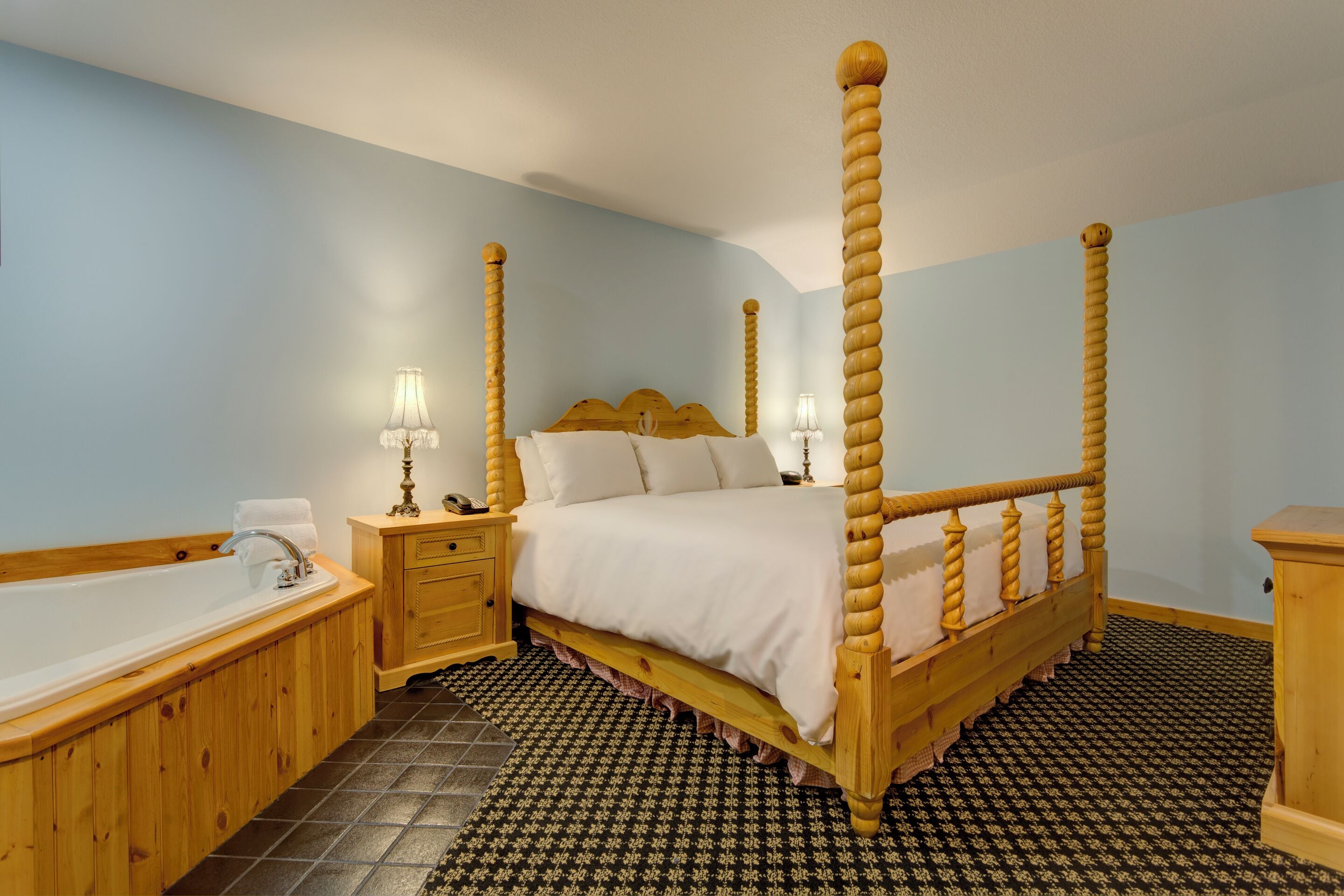 Una cama con dosel, un cabecero de madera, una mesita de noche con lámpara, un banco de madera y una bañera.