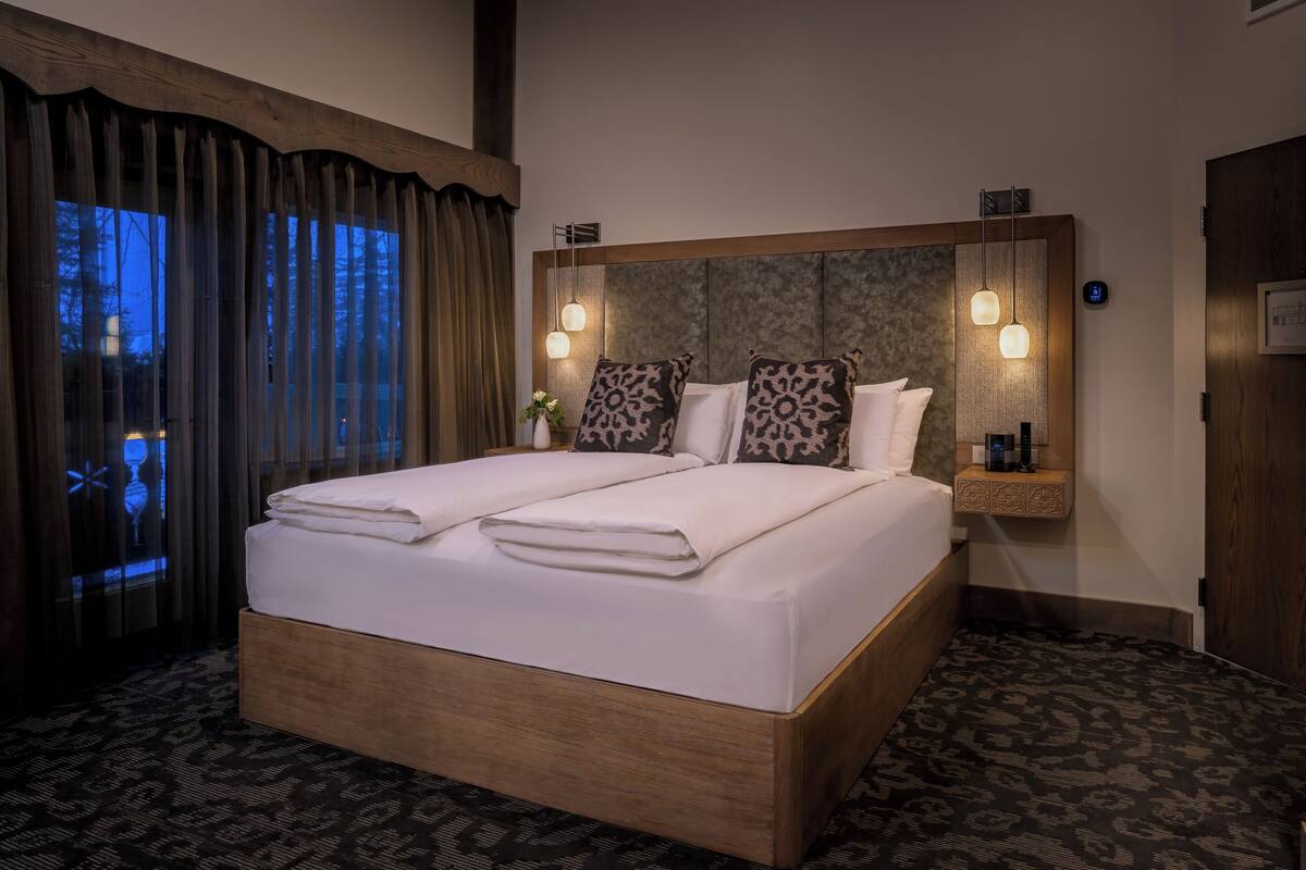 A double bed with a wooden headboard, two bedside tables with lamps, a view of the outdoors through a window with curtains, and a patterned carpet.