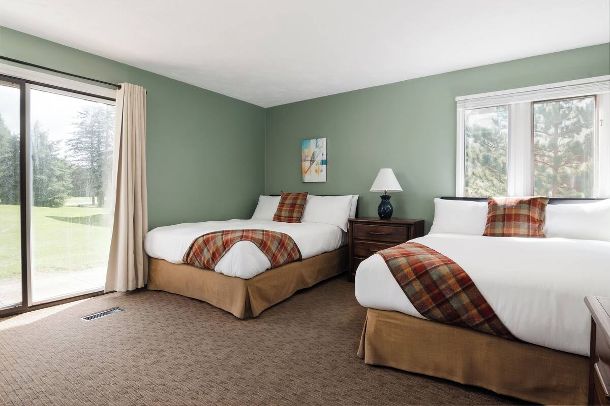 A hotel room with two beds, a window, a painting, and a lamp.
