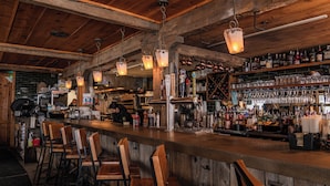 Un espace bar doté de tabourets en bois, de luminaires suspendus et d’étagères remplies de bouteilles et de verres.