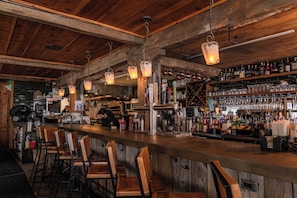 Uma área de bar com banquetas de madeira, luminárias pendentes e prateleiras repletas de garrafas e copos.