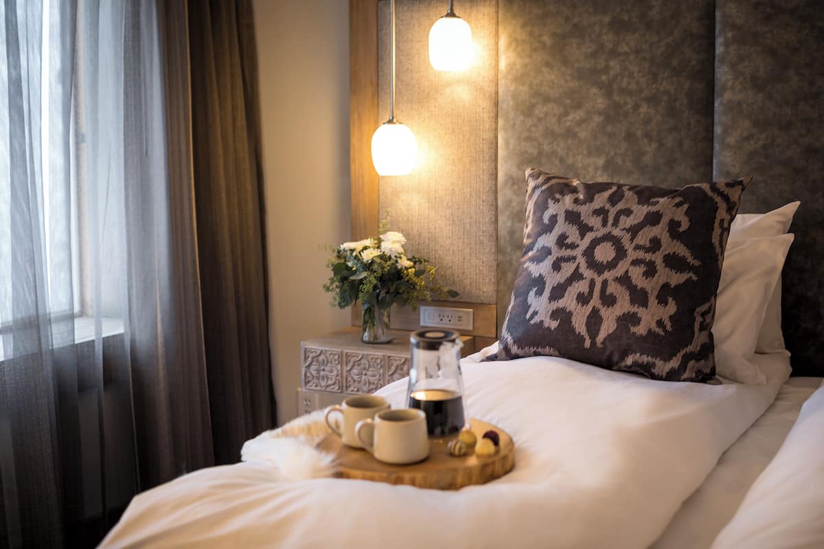 A neatly made bed with a tray of mugs and a coffee pot, a patterned pillow, and a nightstand with a vase of flowers.