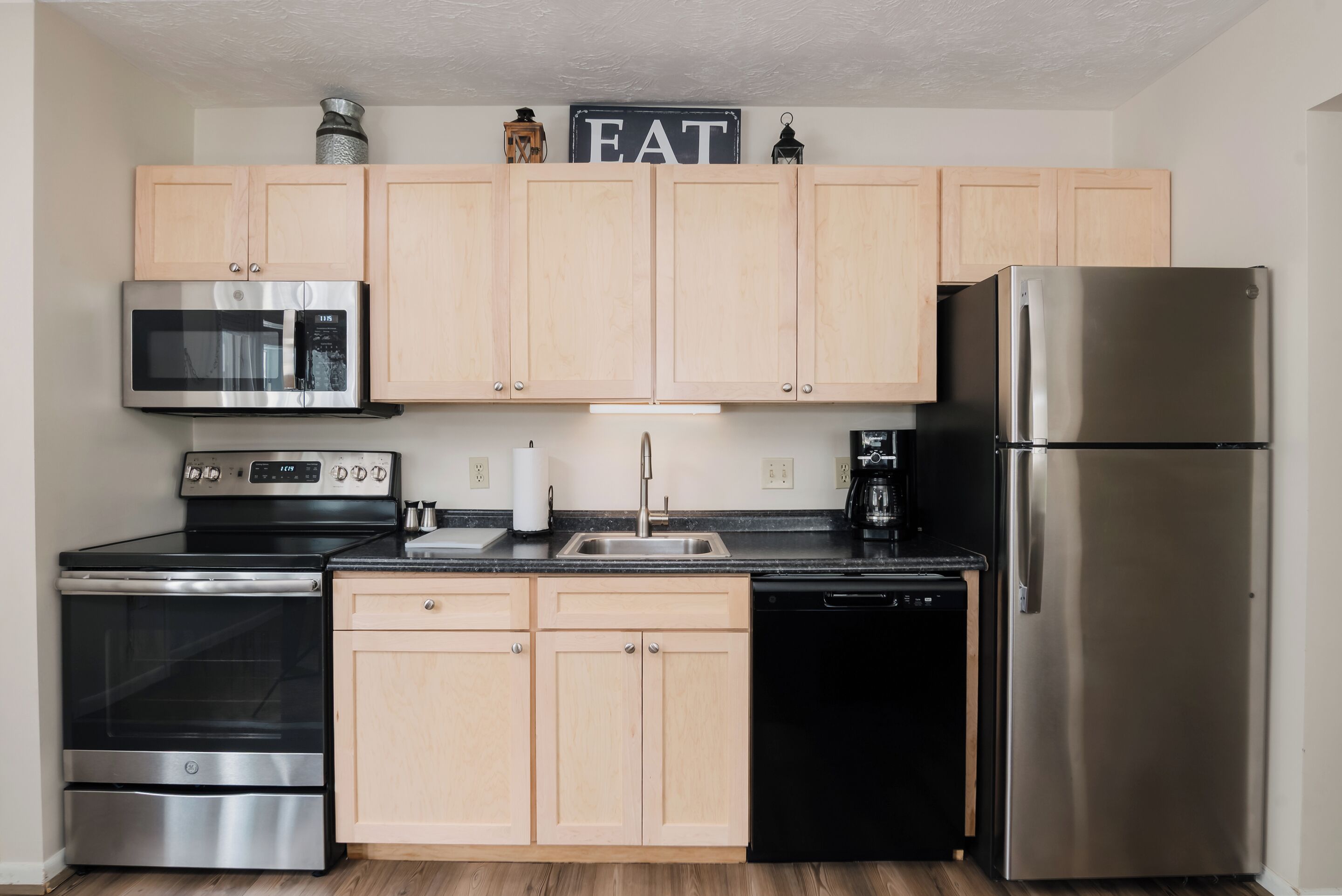 A modern kitchen with light wooden cabinets, stainless steel appliances including a microwave, oven, dishwasher, and refrigerator.