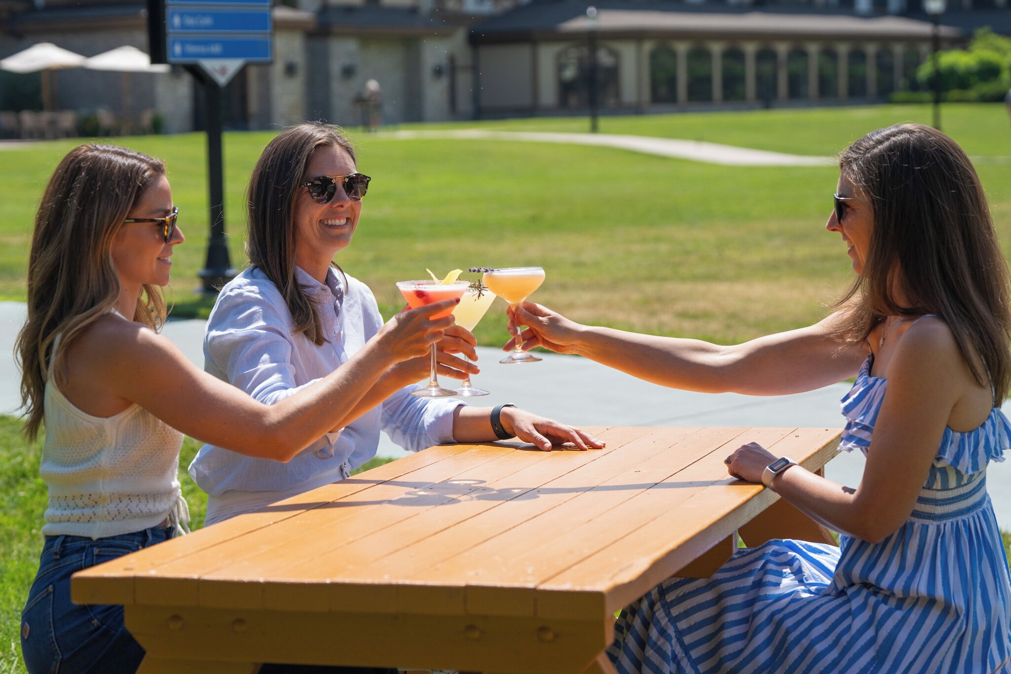 Three people toasting with drinks at a wooden picnic table in a grassy outdoor area with a building in the background.