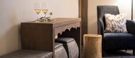 Une petite table d’appoint en bois, sur laquelle reposent deux verres de vin blanc, un banc rembourré, un tabouret rond en bois et un fauteuil sombre agrémenté d’un plaid en fourrure.